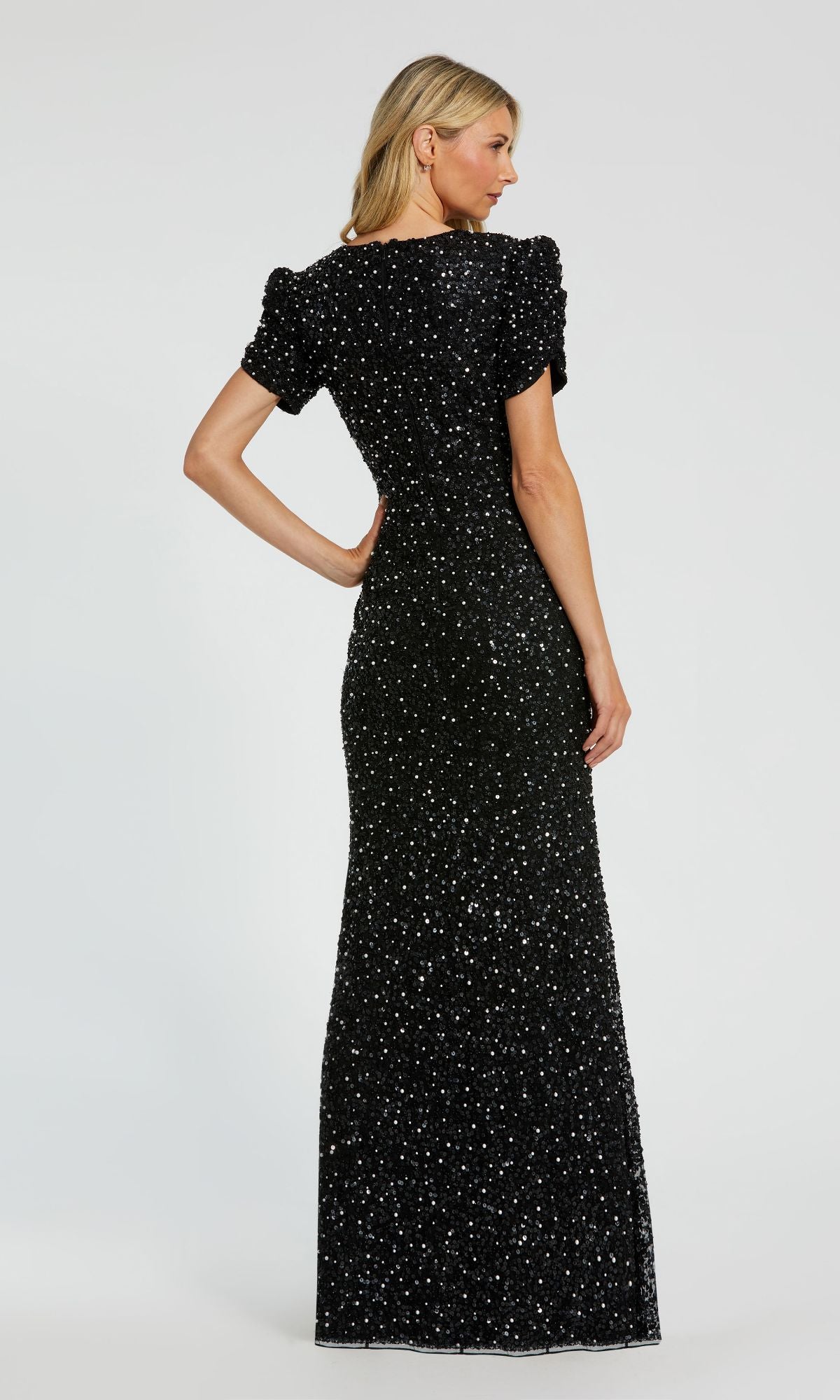 A woman stands facing away, wearing the Mac Duggal 24003 Short Sleeve Sequin Formal Dress—a floor-length black evening gown with sparkling embellishments.